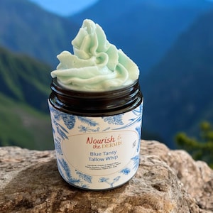 May include: A jar of blue tansy tallow whip with a white label that says "Nourish the Dermis" and "Blue Tansy Tallow Whip". The jar is sitting on a rock with a mountain background.