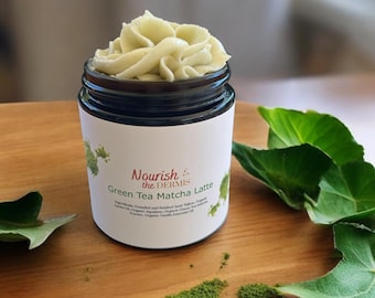 May include: A glass jar with a white label that reads "Nourish the Dermis Green Tea Matcha Latte". The jar is filled with a green, whipped cream-like substance. The jar is sitting on a wooden surface with green leaves and a sprinkle of green powder.