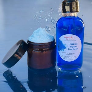 May include: A blue glass bottle with a dropper top and a white label that reads "Nourish the Dermis" and "GHK-Cu Peptide Face Serum". The bottle is sitting on a blue surface with water droplets. A brown glass jar with a white cream inside is also on the surface.