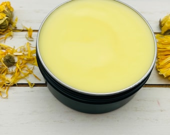 May include: A close-up of a small, round, black container filled with a pale yellow balm. The container is sitting on a white wooden surface with dried yellow flower petals scattered around it.