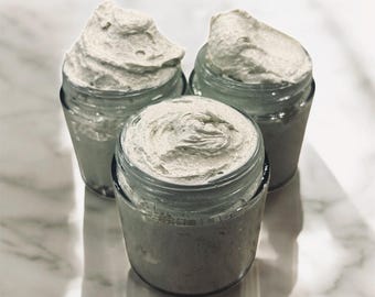 May include: Three small, clear glass jars filled with a light gray, whipped cream-like substance. The jars are arranged on a white marble surface, with the product overflowing the rims.