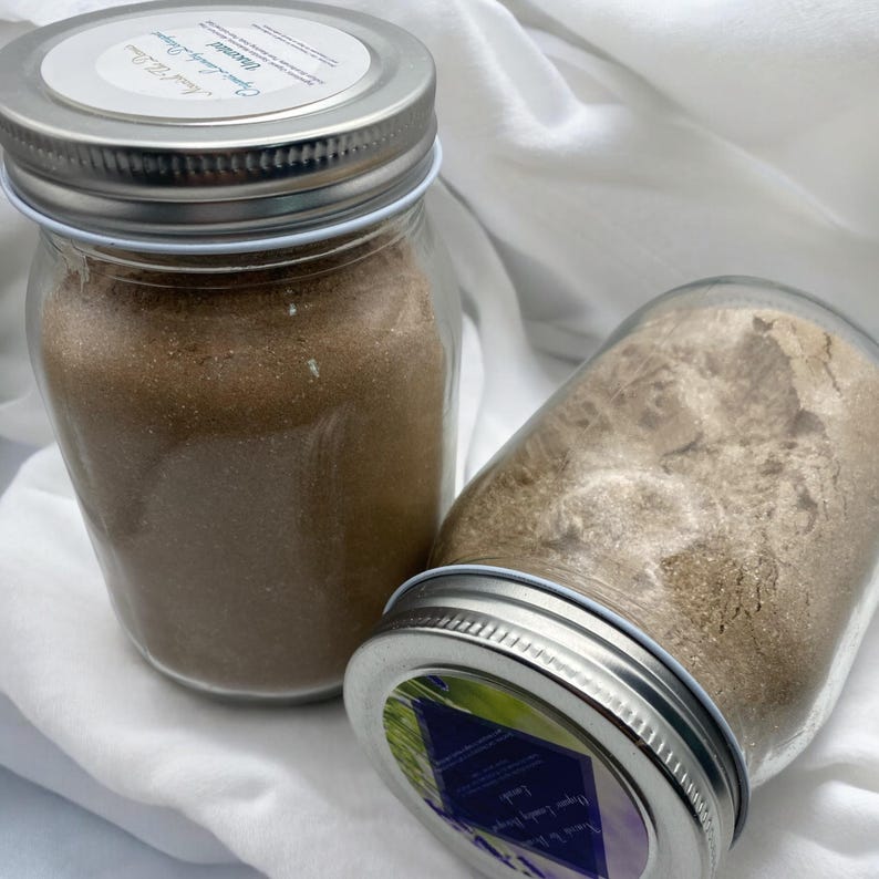 May include: Two clear glass jars with silver lids filled with a brown powder. The jars are labeled with text on a white and blue sticker. The jars are sitting on a white cloth.