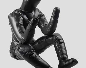 Könnte beinhalten: Eine schwarze, sitzende Figur mit rundem Kopf, erhobenen Armen und ausgestreckten Beinen. Die Figur besteht aus einem glänzenden, gepolsterten Material und befindet sich in einer entspannten Position. Das Gesamtdesign ist minimalistisch und modern.