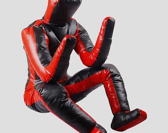 Könnte beinhalten: Eine rot-schwarze Trainingspuppe in sitzender Position. Die Puppe ist so gestaltet, dass sie einer menschlichen Figur ähnelt, mit Kapuze, Armen und Beinen. Das Farbschema ist hauptsächlich rot und schwarz.