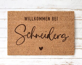 May include: A brown coir doormat with the text "Willkommen bei Schneiders" in black cursive lettering and a small heart.