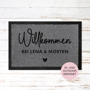 May include: A grey welcome mat with black text that reads "Willkommen bei Lena & Morten" with a small black heart below the text. The mat has a black border and a pink circle with white text that reads "IN-UND OUTDOOR GEEIGNET".
