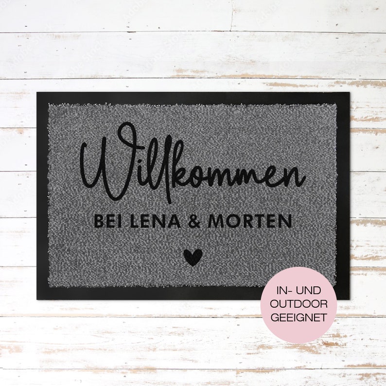 May include: A grey welcome mat with black text that reads "Willkommen bei Lena & Morten" with a small black heart below the text. The mat has a black border and a pink circle with white text that reads "IN-UND OUTDOOR GEEIGNET".