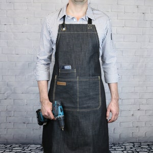 May include: A black denim apron with a large pocket on the front, a smaller pocket on the left side, and two D-rings at the top. The apron has yellow stitching and a brown leather patch on the front. A person is wearing the apron and holding a cordless drill.