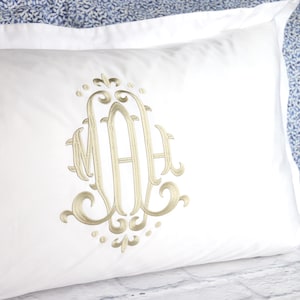May include: A rectangular white pillowcase featuring a centered, ornate gold-colored embroidered monogram with the letters MAH. The embroidery displays intricate swirling flourishes and decorative scrollwork details surrounding the central letters.