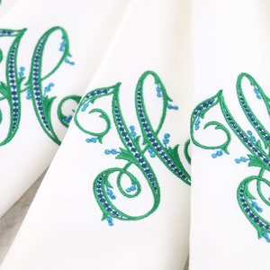 May include: Set of white linen napkins with green and blue embroidered floral monograms. The monogram is a stylized letter H.