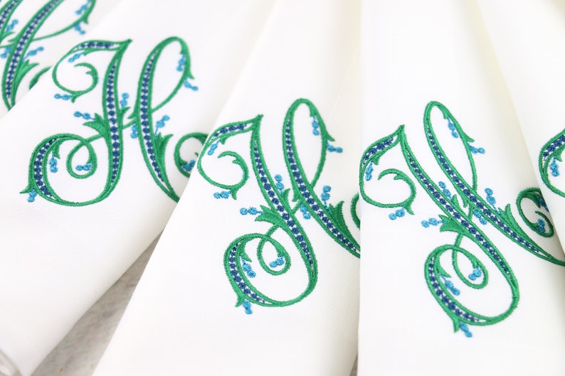 May include: Set of white linen napkins with green and blue embroidered floral monograms. The monogram is a stylized letter H.