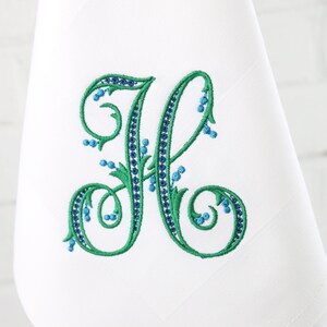 May include: A white embroidered kitchen towel with a green and blue floral monogram of the letter "H".