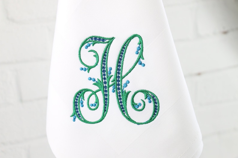 May include: A white embroidered kitchen towel with a green and blue floral monogram of the letter "H".