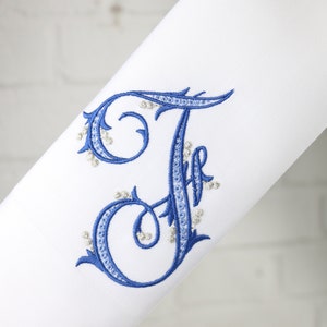 May include: A white fabric with a blue embroidered letter "F" with silver beads around the edges.