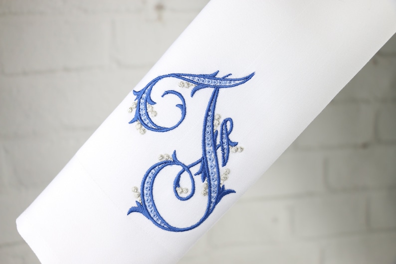 May include: A white fabric with a blue embroidered letter "F" with silver beads around the edges.