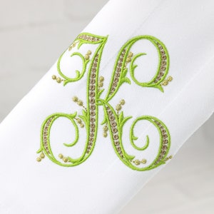 May include: A white fabric napkin with a green embroidered letter K. The letter is in a script font with decorative flourishes and is outlined with a gold thread.