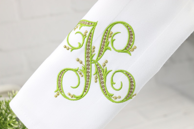 May include: A white fabric napkin with a green embroidered letter K. The letter is in a script font with decorative flourishes and is outlined with a gold thread.