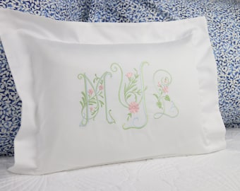 May include: White pillowcase with embroidered floral initials in green and pink. The pillowcase has a ruffled edge and is placed on a white bedspread. A blue patterned pillow is in the background.