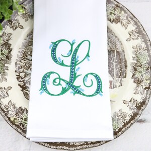 May include: White linen napkin with a green embroidered letter L with blue accents. The letter is surrounded by a leafy design.
