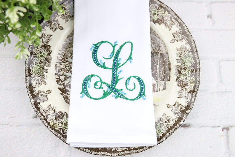 May include: White linen napkin with a green embroidered letter L with blue accents. The letter is surrounded by a leafy design.