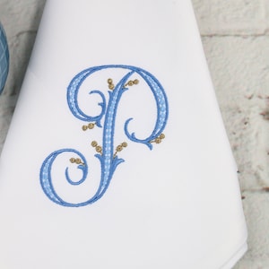 May include: A white fabric napkin with a blue embroidered letter "P" with gold accents. The letter is in a decorative script font with swirls and flourishes.