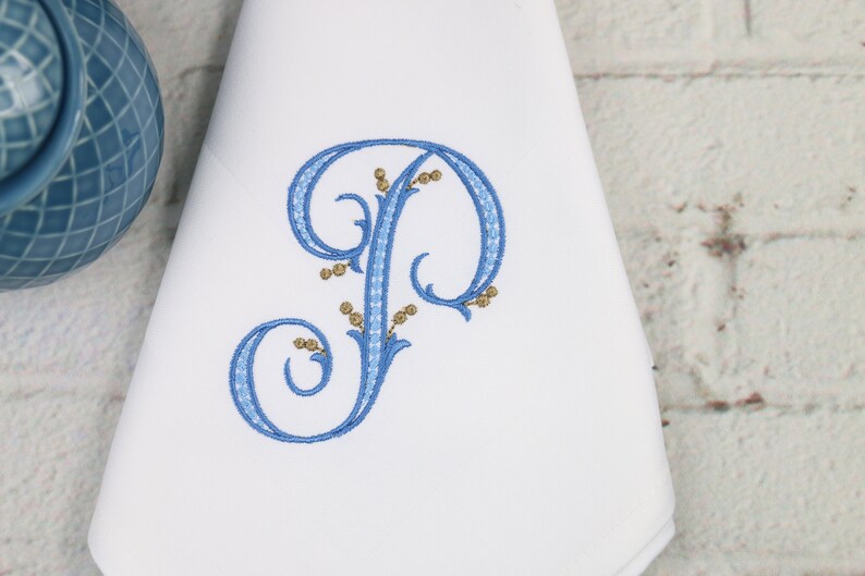 May include: A white fabric napkin with a blue embroidered letter "P" with gold accents. The letter is in a decorative script font with swirls and flourishes.