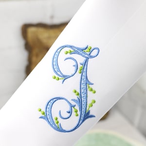 May include: A white fabric with a blue and green embroidered letter J. The letter is in a script font with decorative flourishes and green leaves.