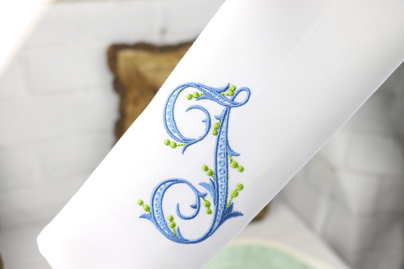 May include: A white fabric with a blue and green embroidered letter J. The letter is in a script font with decorative flourishes and green leaves.