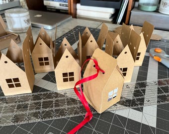 May include: Kraft paper house-shaped gift boxes with square windows and a red ribbon. The boxes are arranged on a cutting mat with a ruler and rotary cutter. The boxes are ready for assembly.