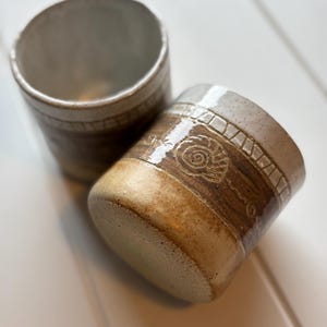 Könnte beinhalten: Zwei zylindrische Keramikbecher mit einer gesprenkelten cremefarbenen Glasur am oberen Rand und einem strukturierten braunen Band in der Mitte. Der braune Bereich ist mit einem geprägten Spiralmuschel-Motiv und einem geometrischen Randmuster verziert.