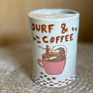 May include: A cylindrical ceramic mug with a speckled light grey finish. The front features the brown text SURF & COFFEE above a painted illustration of a person surfing on a wave inside a pink coffee cup. A brown and white chequered pattern borders the base.