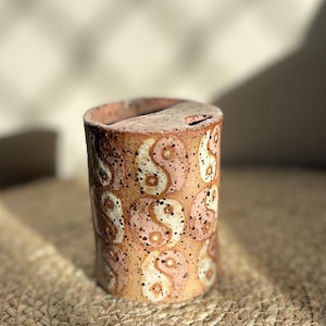 May include: A cylindrical ceramic coin bank with a speckled tan and brown finish. The exterior is decorated with a repeating pattern of yin and yang symbols in shades of cream and light pink. A narrow coin slot is visible on the top surface.