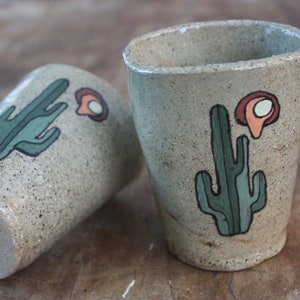 May include: Two ceramic mugs with a speckled beige glaze. Each mug features a hand-painted green cactus with a red and orange flower.