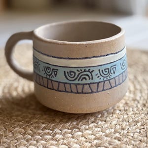 May include: A beige ceramic mug with a rounded shape and a single handle. The exterior features a horizontal band decorated with light blue and brown geometric patterns, including stylised swirls and triangles outlined in dark brown ink.