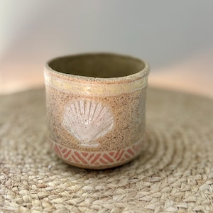 May include: A speckled ceramic cup featuring a light pink embossed seashell design on the front. The rim has a smooth cream-coloured band, and the base is decorated with a repeating geometric pattern in a muted reddish-orange hue.