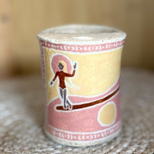 May include: A cylindrical ceramic cup with a speckled off-white base. The exterior features a hand-painted design with a yellow background, a pink wave-like shape, and a figure in a dark red leotard. Decorative borders with white dots frame the top and bottom.