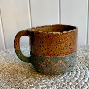 May include: A ceramic mug with a speckled brown upper section and a green lower section featuring a carved mountain range design. The handle is attached to the side and is finished in the same speckled green glaze as the base of the mug.