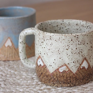 May include: Two ceramic mugs with mountain designs. One mug is speckled with a cream base and brown speckles, and the other is a light blue. Both mugs have a mountain range design in brown and white.