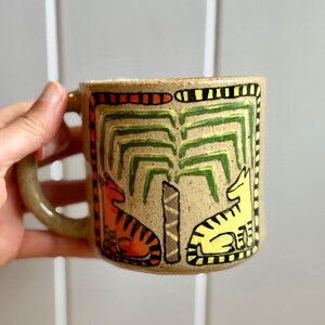 May include: A speckled beige ceramic mug featuring a hand-painted design of a central palm tree flanked by an orange tiger on the left and a yellow tiger on the right, all enclosed within a rectangular border of orange and black stripes.