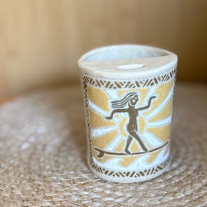 May include: A cylindrical ceramic wall planter with a textured off-white finish. The front features a hand-painted graphic of a figure on a surfboard against a yellow sunburst and white waves, framed by a geometric border pattern.