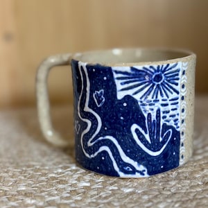 May include: A ceramic mug with a speckled beige base and a dark blue painted panel. The blue section features a white line drawing of a winding snake, a sun with rays, and a cactus. A vertical border of white circles decorates the right side.