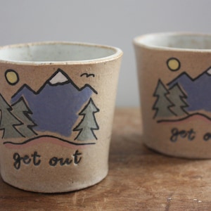Puede incluir: Dos tazas de cerámica con una escena de montaña y las palabras "get out" en la parte inferior. Las tazas son beige con acentos marrones, azules y amarillos.