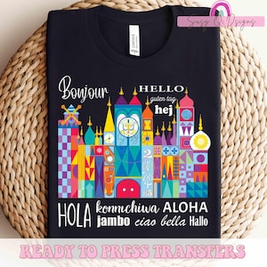 Op de afbeelding: Zwart T-shirt met een kleurrijke afbeelding van een kasteel met verschillende talen erop, waaronder Bonjour, Hello, Guten Tag, Hej, Hola, Konnichiwa, Aloha, Jambo, Ciao, Bella en Hallo. De tekst "READY TO PRESS TRANSFERS" staat onderaan het shirt.