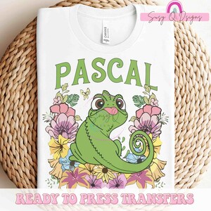 May include: White t-shirt with a green chameleon cartoon character named Pascal surrounded by colorful flowers. The text "PASCAL" is above the character. The text "READY TO PRESS TRANSFERS" is at the bottom of the shirt.