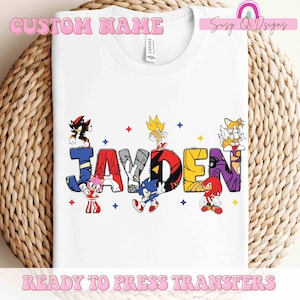 May include: White t-shirt with a custom name design featuring characters from the Sonic the Hedgehog video game series. The name "JAYDEN" is spelled out with each letter formed by a different character. The characters are in their signature colors and poses. The shirt is ready to press.