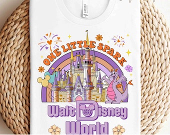 Könnte beinhalten: Ein weißes T-Shirt mit einem lila, orange und rosa Design, das eine Zeichentrickburg, Feuerwerk und einen Drachen zeigt. Der Text "One Little Spark" ist in einem lila Kreis über der Burg geschrieben. Der Text "Walt Disney World" ist unter der Burg geschrieben. Der Text "Ready to Press Transfers" ist in rosa am unteren Rand des Shirts geschrieben.