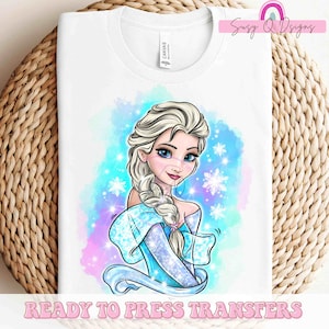 May include: White t-shirt featuring a cartoon illustration of a princess with blonde hair, blue eyes, and a blue dress. The design includes snowflakes and a watercolor background in shades of blue, purple, and pink. Text at the bottom reads "READY TO PRESS TRANSFERS".
