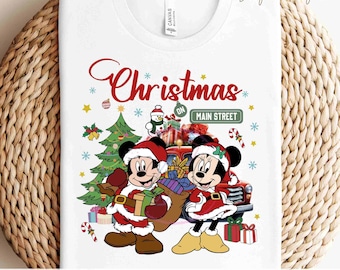 Puede incluir: Una camiseta blanca con un diseño navideño rojo y blanco con Mickey y Minnie Mouse vestidos de Papá Noel. El diseño incluye un árbol de Navidad, regalos y un camión rojo con el texto "Christmas on Main Street".