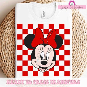 May include: White t-shirt featuring a Minnie Mouse graphic. The design includes Minnie's face with a red bow, set against a red and white checkered background. The words "READY TO PRESS TRANSFERS" are printed at the bottom.