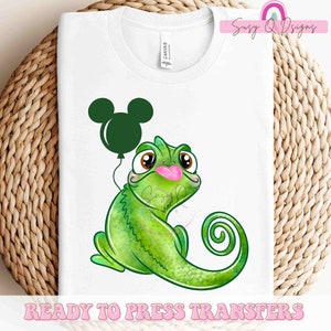 May include: A green chameleon with a pink tongue holding a green Mickey Mouse balloon.  The chameleon is cartoon-style and has large eyes.  The image is on a white t-shirt.  Ready to Press Transfers.
